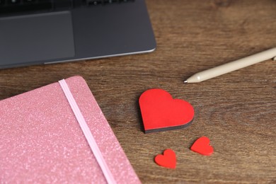 Photo of Decorative hearts, laptop, notebook and pen on wooden table, closeup. Online dating site