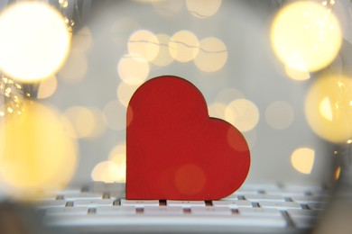 Photo of Online dating site. Wooden heart on computer keyboard against blurred lights, view through magnifying glass. Bokeh effect
