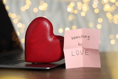 Photo of Online dating site. Sticky note with text Searching For Love, decorative heart and laptop on wooden table, closeup. Bokeh effect