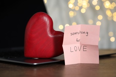 Photo of Online dating site. Sticky note with text Searching For Love, decorative heart and laptop on wooden table, closeup. Bokeh effect