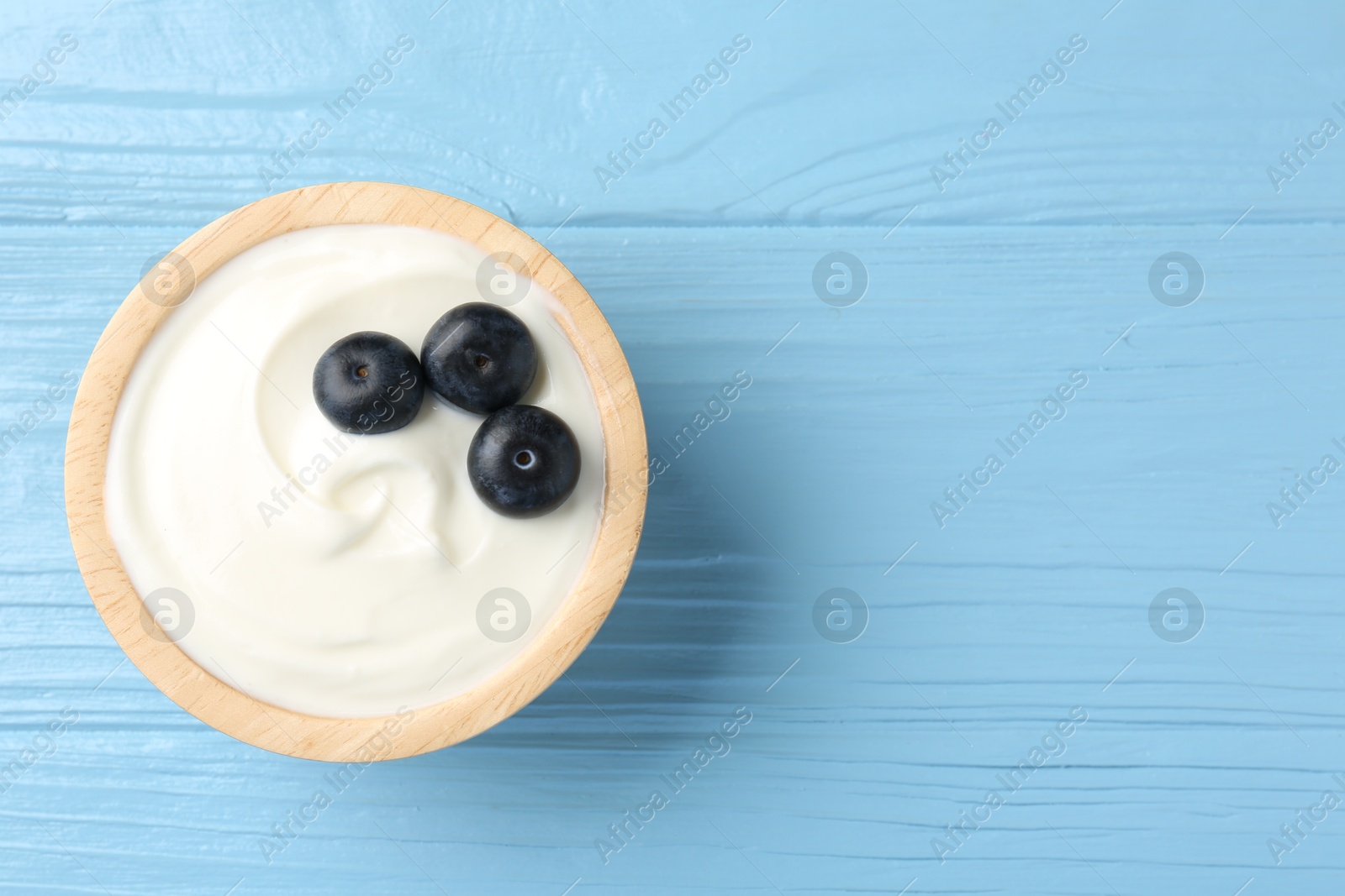 Photo of Tasty yoghurt with blueberries in bowl on light blue wooden table, top view. Space for text