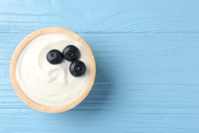 Photo of Tasty yoghurt with blueberries in bowl on light blue wooden table, top view. Space for text