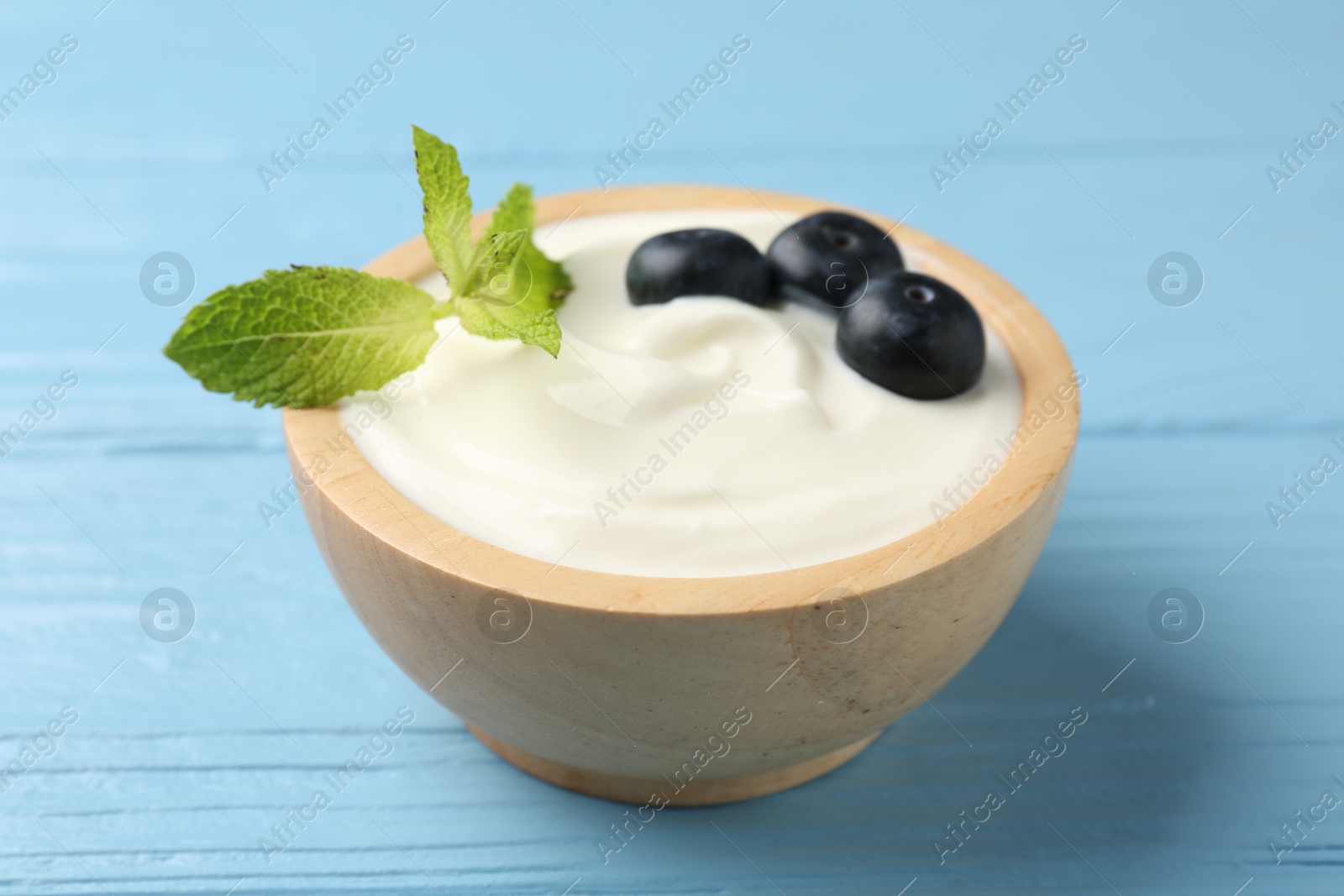 Photo of Tasty yoghurt with blueberries and mint in bowl on light blue wooden table, closeup