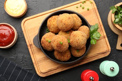 Photo of Delicious fried croquette balls with green onion, parsley, sauces and spices on dark textured table, flat lay