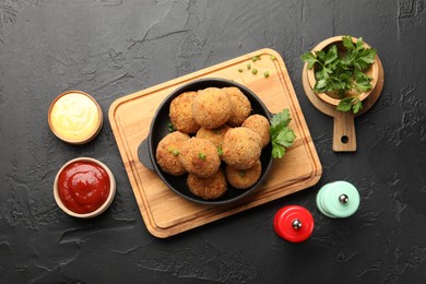 Photo of Delicious fried croquette balls with green onion, parsley, sauces and spices on dark textured table, flat lay