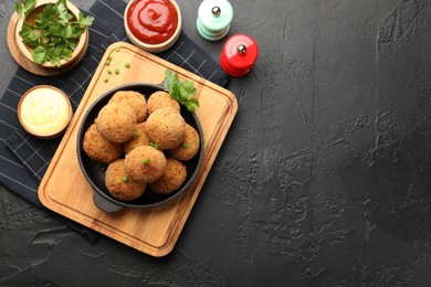 Photo of Delicious fried croquette balls with green onion, parsley, sauces and spices on dark textured table, flat lay. Space for text
