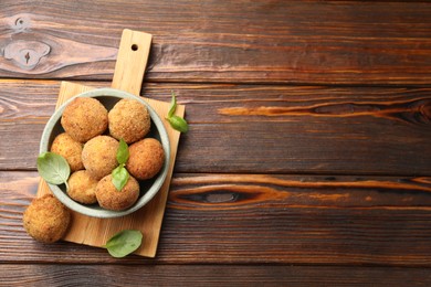 Photo of Delicious fried croquette balls with basil in bowl on wooden table, top view. Space for text