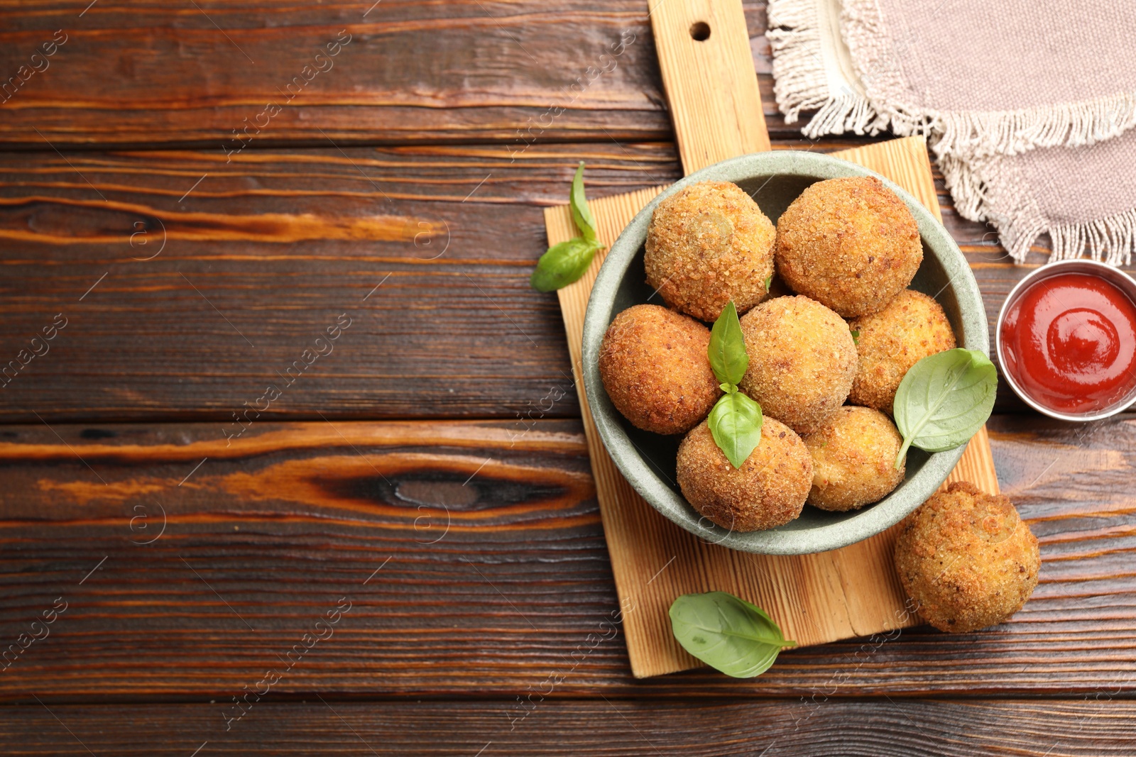 Photo of Delicious fried croquette balls with basil in bowl and ketchup on wooden table, flat lay. Space for text