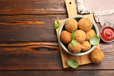 Photo of Delicious fried croquette balls with basil in bowl and ketchup on wooden table, flat lay. Space for text