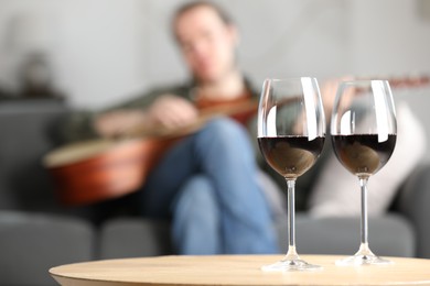 Photo of Glasses with red wine on table and man playing guitar indoors, selective focus
