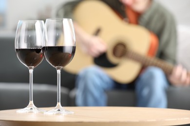 Photo of Glasses with red wine on table and man playing guitar indoors, selective focus