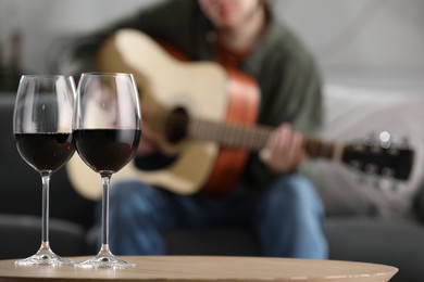 Photo of Glasses with red wine on table and man playing guitar indoors, selective focus