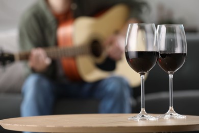 Photo of Glasses with red wine on table and man playing guitar indoors, selective focus