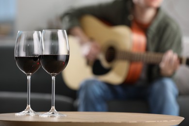 Photo of Glasses with red wine on table and man playing guitar indoors, selective focus