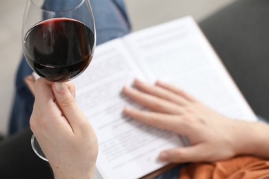 Photo of Woman with glass of wine reading book on sofa at home, closeup