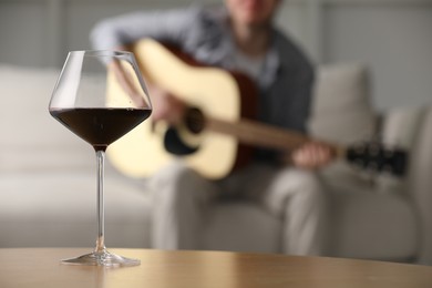 Photo of Glass with red wine on table and man playing guitar indoors, selective focus