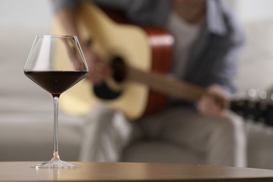 Photo of Glass with red wine on table and man playing guitar indoors, selective focus