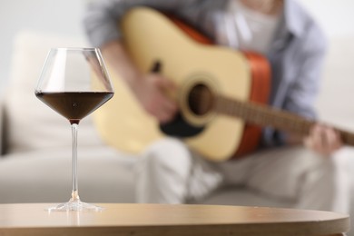 Photo of Glass with red wine on table and man playing guitar indoors, selective focus