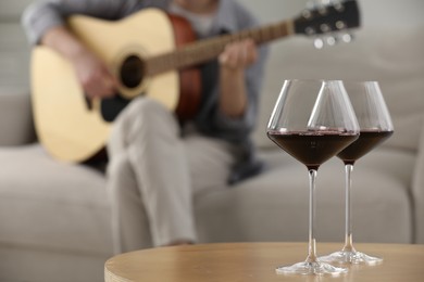 Photo of Glasses with red wine on table and man playing guitar indoors, selective focus