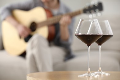 Photo of Glasses with red wine on table and man playing guitar indoors, selective focus