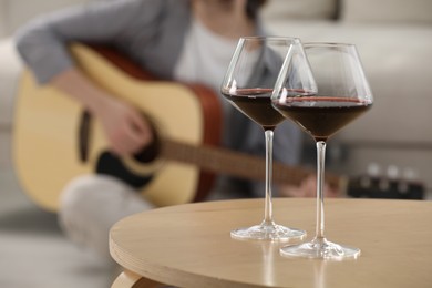Photo of Glasses with red wine on table and man playing guitar indoors, selective focus