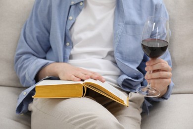 Photo of Woman with glass of wine reading book on sofa at home, closeup