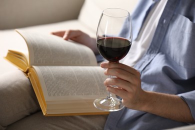 Photo of Woman with glass of wine reading book on sofa at home, closeup