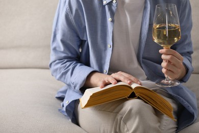 Photo of Woman with glass of wine reading book on sofa at home, closeup