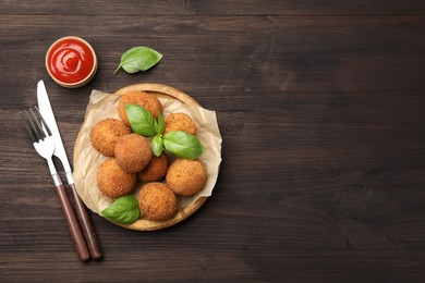 Photo of Delicious fried croquette balls with basil, cutlery and ketchup on wooden table, flat lay. Space for text