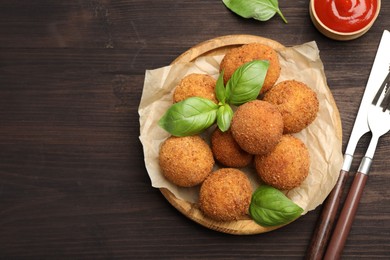 Photo of Delicious fried croquette balls with basil, cutlery and ketchup on wooden table, flat lay. Space for text