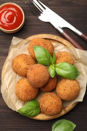 Photo of Delicious fried croquette balls with basil, cutlery and ketchup on wooden table, flat lay