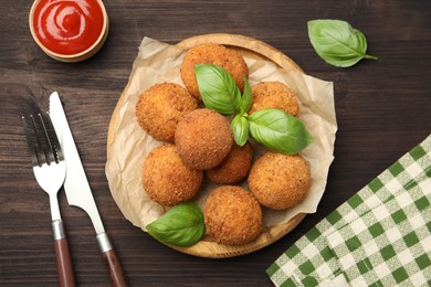 Photo of Delicious fried croquette balls with basil, cutlery and ketchup on wooden table, flat lay