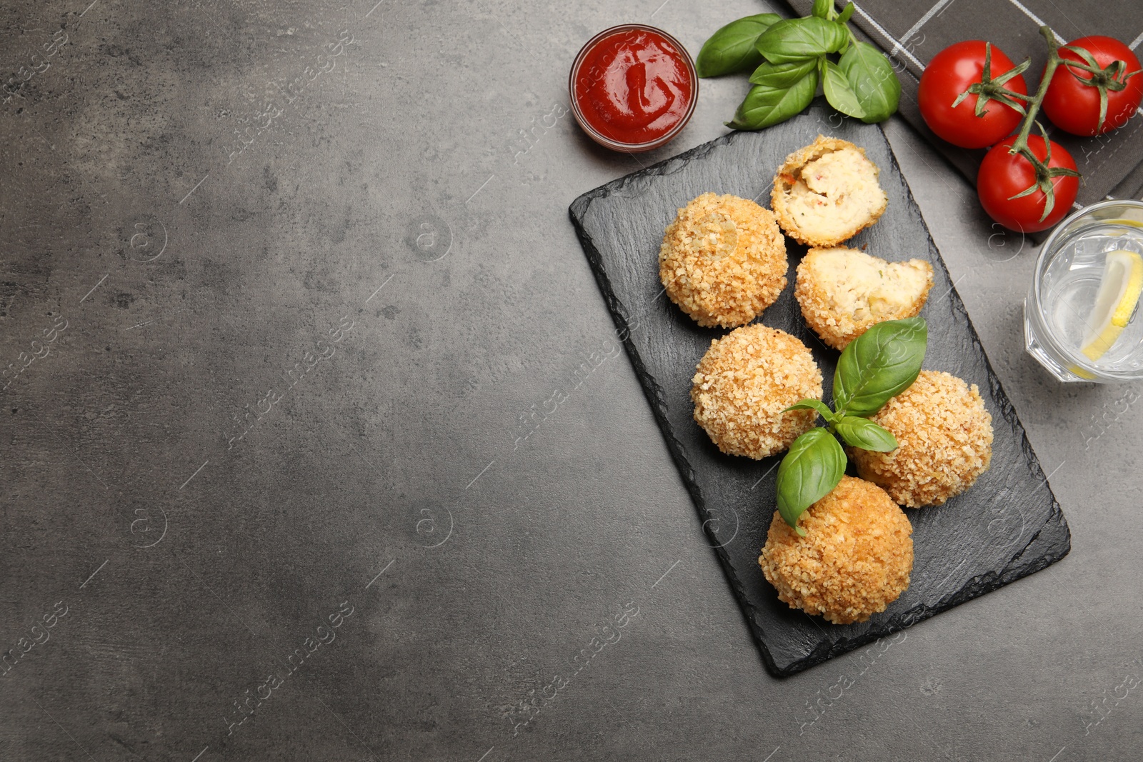 Photo of Delicious fried croquette balls with basil, tomato, water and ketchup on grey textured table, flat lay. Space for text