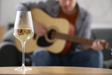 Photo of Glass with white wine on table and man playing guitar indoors, selective focus