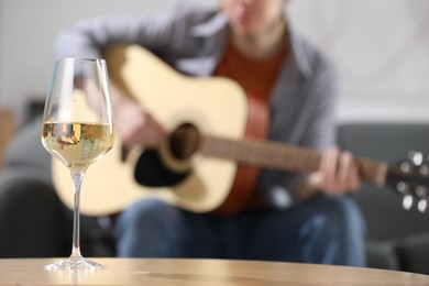 Photo of Glass with white wine on table and man playing guitar indoors, selective focus