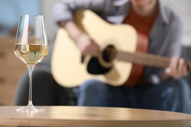 Photo of Glass with white wine on table and man playing guitar indoors, selective focus