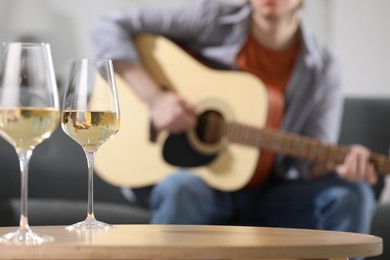 Photo of Glasses with white wine on table and man playing guitar indoors, selective focus