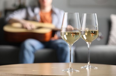 Photo of Glasses with white wine on table and man playing guitar indoors, selective focus