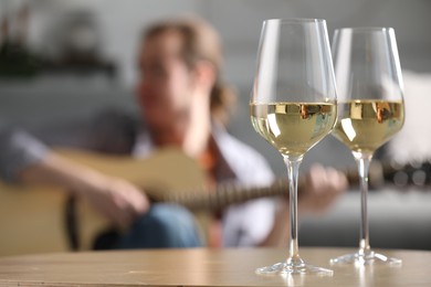 Photo of Glasses with white wine on table and man playing guitar indoors, selective focus