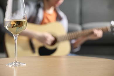 Photo of Glass with white wine on table and man playing guitar indoors, selective focus