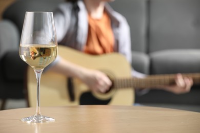 Photo of Glass with white wine on table and man playing guitar indoors, selective focus