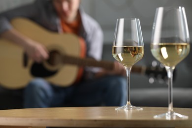 Photo of Glasses with white wine on table and man playing guitar indoors, selective focus