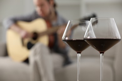 Photo of Glasses with red wine and man playing guitar indoors, selective focus