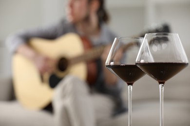 Photo of Glasses with red wine and man playing guitar indoors, selective focus