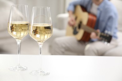 Photo of Glasses with white wine on table and man playing guitar indoors, selective focus