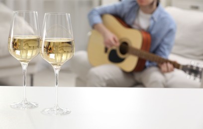Photo of Glasses with white wine on table and man playing guitar indoors, selective focus