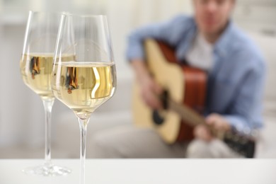 Photo of Glasses with white wine on table and man playing guitar indoors, selective focus