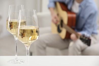 Photo of Glasses with white wine on table and man playing guitar indoors, selective focus