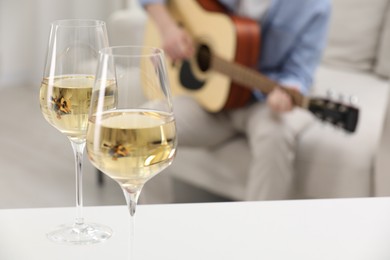 Photo of Glasses with white wine on table and man playing guitar indoors, selective focus