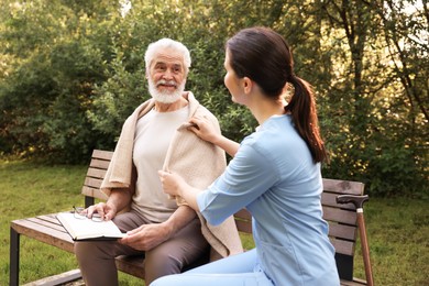 Photo of Nurse supporting smiling elderly man with book and glasses on bench outdoors